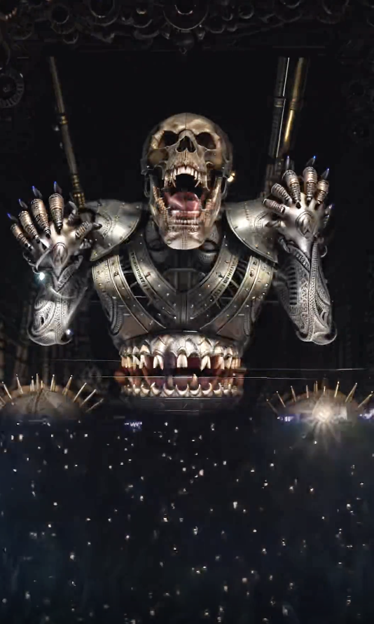 A large mechanical skull with metallic arms is displayed above a crowd of people, with lights illuminating the scene, at a concert or event