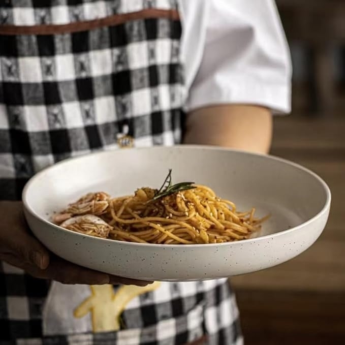 Person in a checkered apron holding a bowl of spaghetti with shrimp and garnished herbs in a white dish