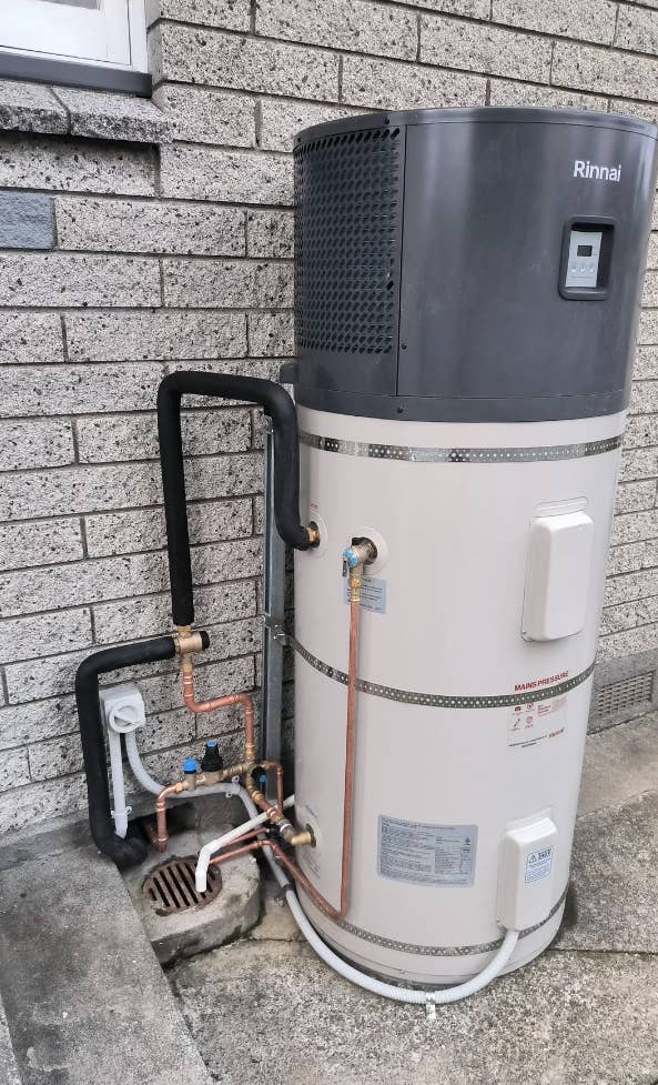 Screenshot of a Reddit post on the Personal Finance NZ subreddit discussing heat pump water heater installation. The heater is next to a brick wall with piping visible