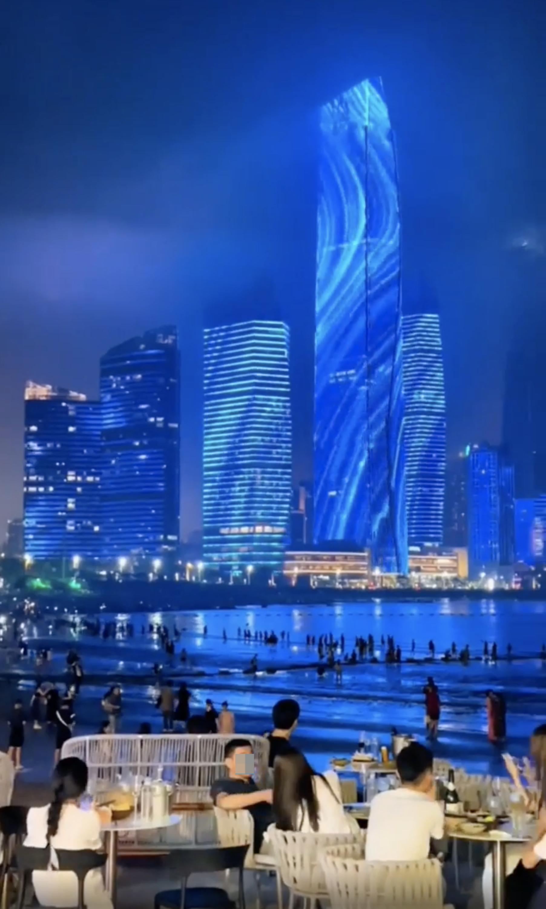 People dining at a beachfront restaurant during nighttime, with tall, illuminated skyscrapers in the background and many people walking along the shoreline