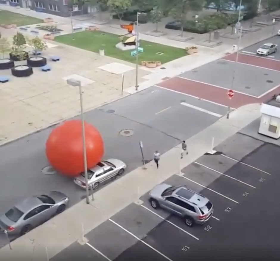 A large red inflatable ball is seen rolling down a city street, with parked cars and a few people walking nearby