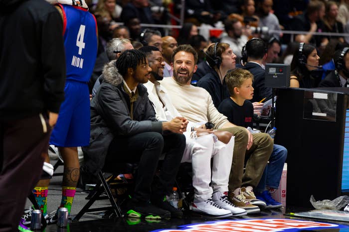 LaMelo Ball and Ben Affleck chat and laugh courtside at an NBA game, with a young boy seated next to Affleck