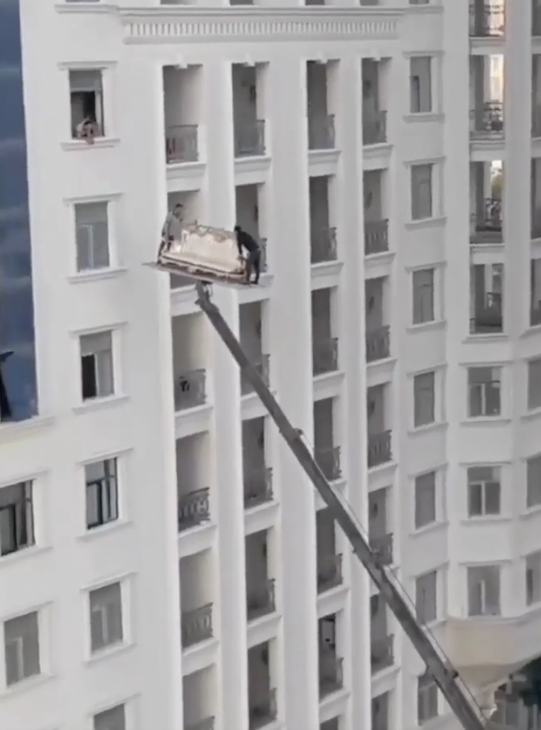 A view of a tall white apartment building with a high-reach crane lifting what appears to be a sofa. Two people on the crane's platform guide it towards a balcony