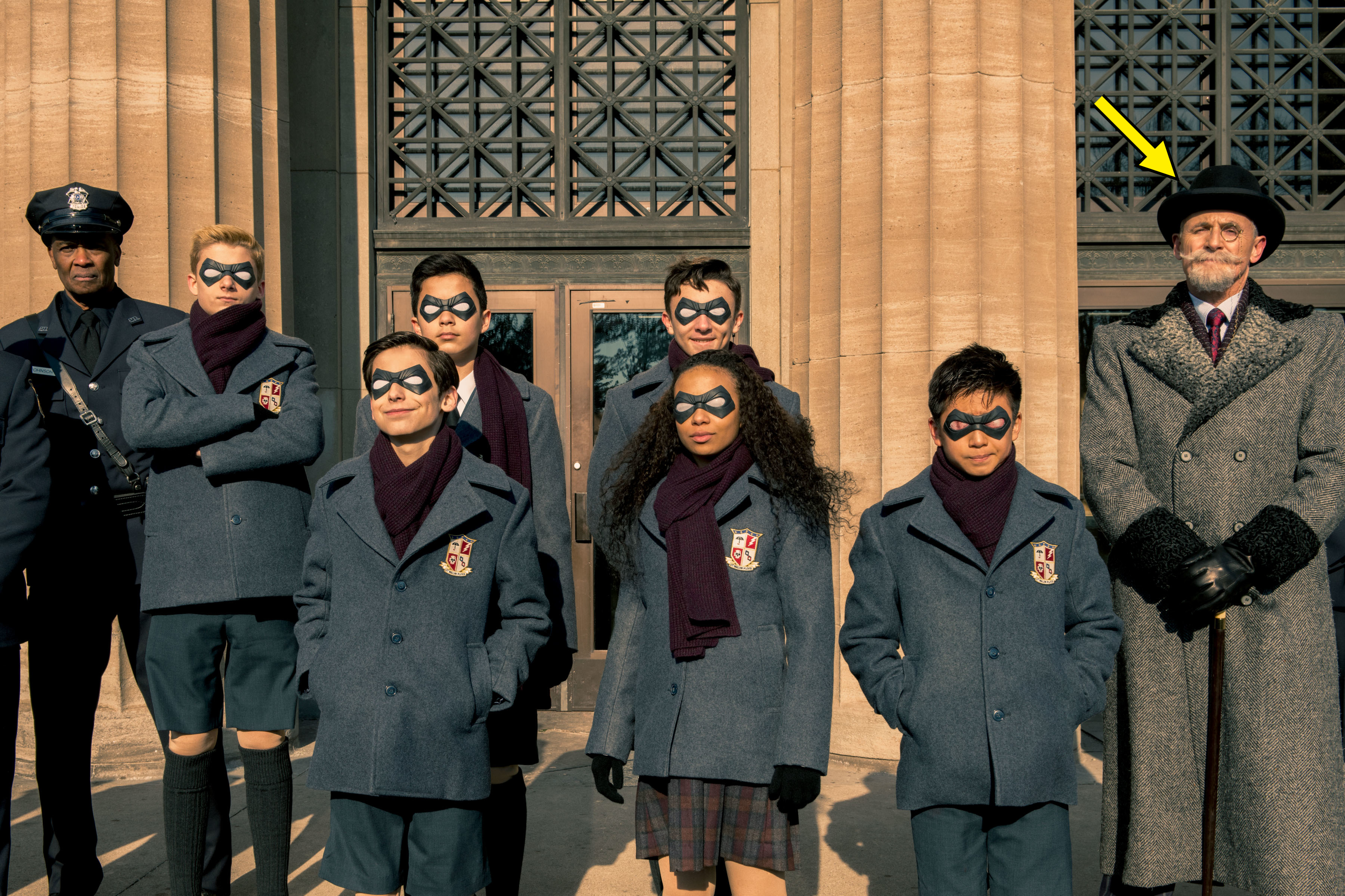 An officer, Cameron Brodeur, Aidan Gallagher, Blake Talabis, Dante Albidone, Eden Cupid, Ethan Hwang, and Colm Feore standing outside a building wearing school uniforms and superhero eye masks in "The Umbrella Academy"