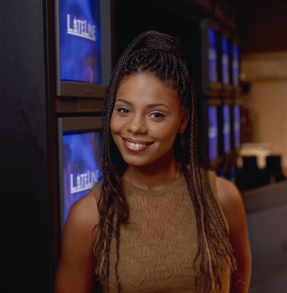 I don't know who the person is. They are standing indoors near several screens displaying the word &quot;LATELINE,&quot; smiling and wearing a sleeveless, textured top