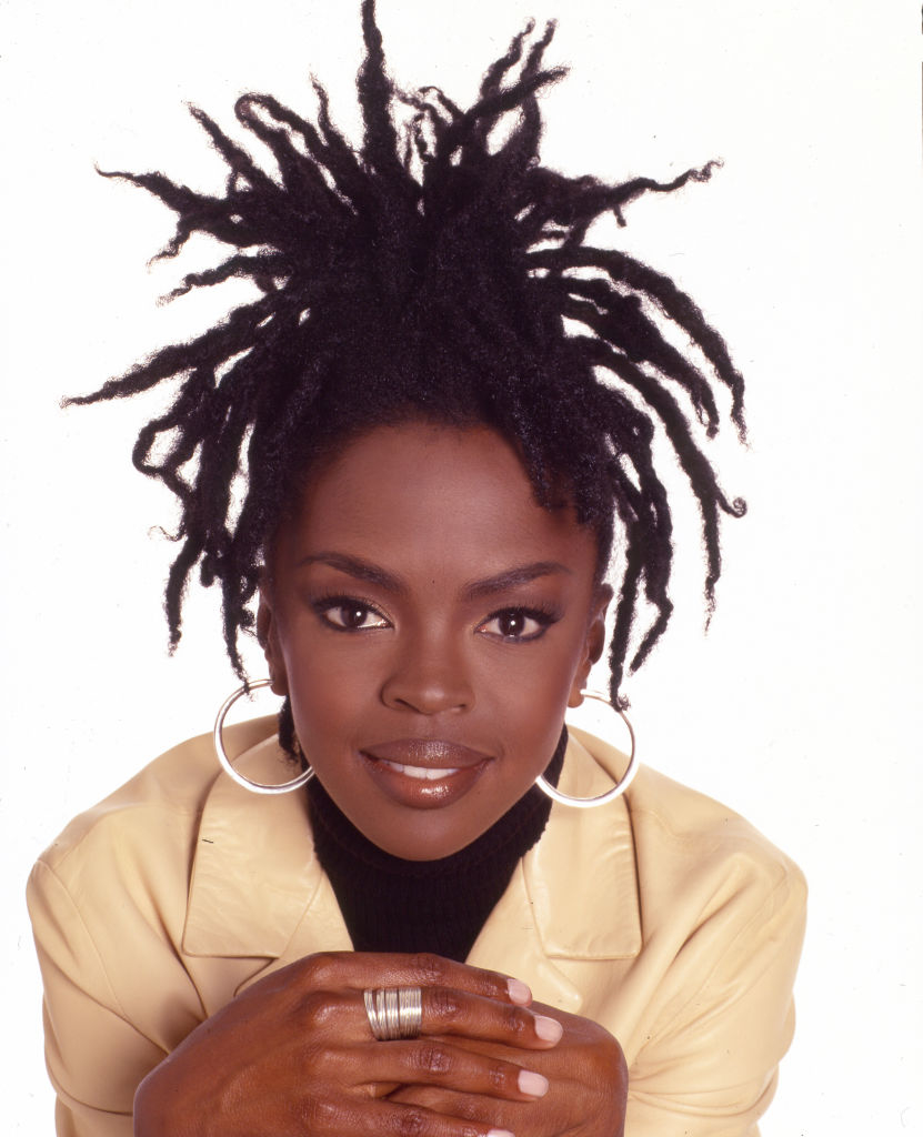 Lauryn Hill poses with a confident smile, wearing a light jacket, black turtleneck, silver hoop earrings, and a silver ring, hands folded under her chin