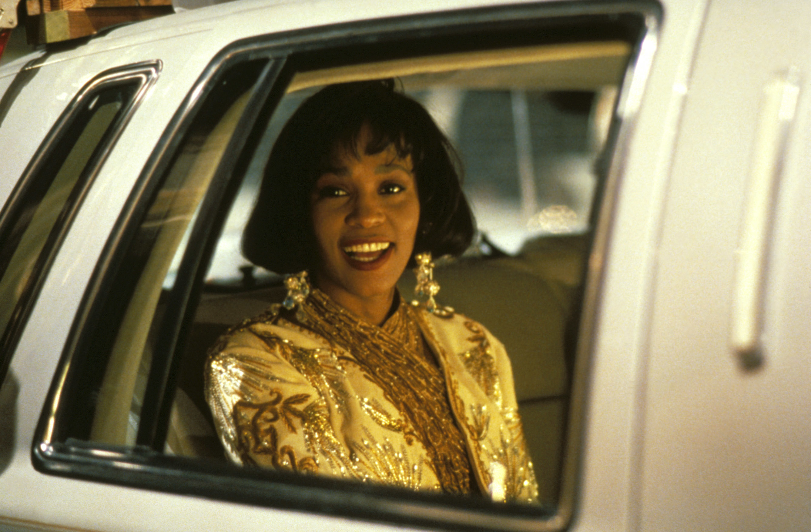 Whitney Houston sits in a car window, smiling, wearing an ornate, embellished outfit and large earrings