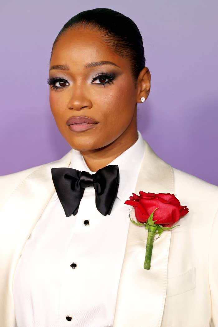 Keke Palmer is wearing a white tuxedo with a black bow tie and a red rose on the lapel, posing on a red carpet