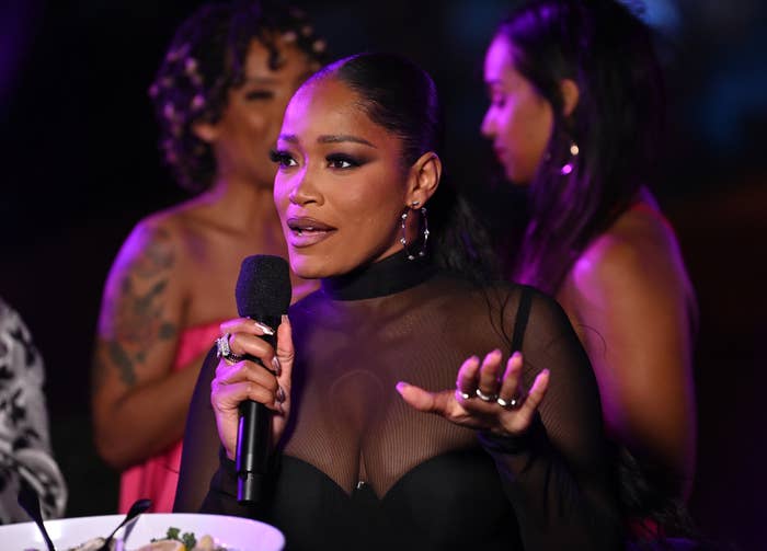 Keke Palmer speaks into a microphone at an event, wearing a sheer black top and large hoop earrings. Two other women are blurred in the background