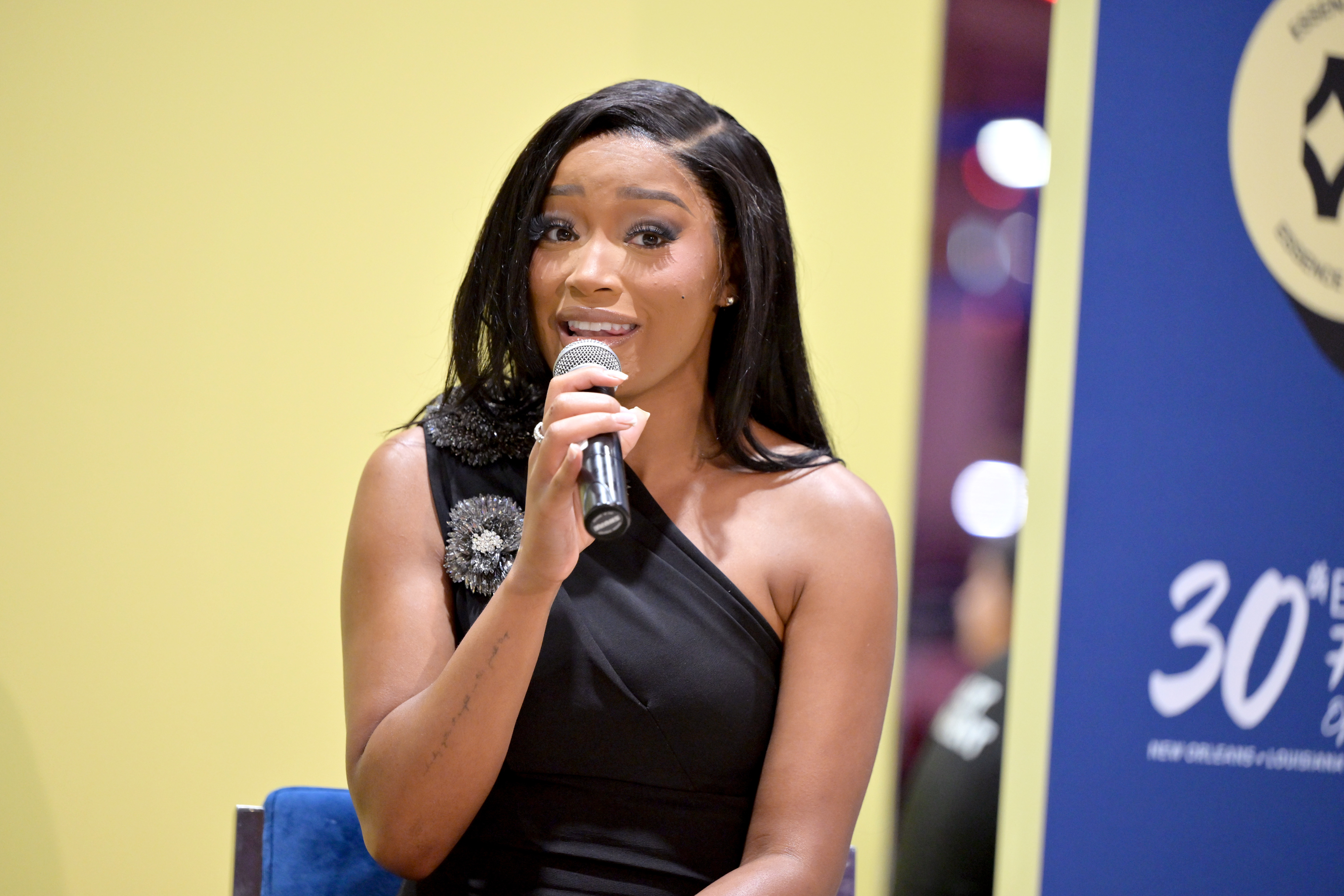 Keke Palmer speaking into a microphone while wearing a stylish, one-shoulder black dress with a decorative brooch on the right shoulder at a public event
