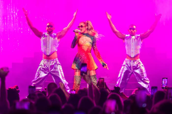 Paris Hilton performs on stage in a colorful outfit with two backup dancers in shiny sleeveless tops and pants. The background is vibrant and lively