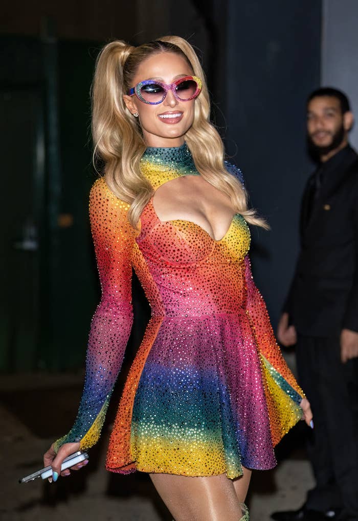 Paris Hilton in a sparkly, rainbow mini-dress and pink sunglasses, holding a marker, smiling at an event