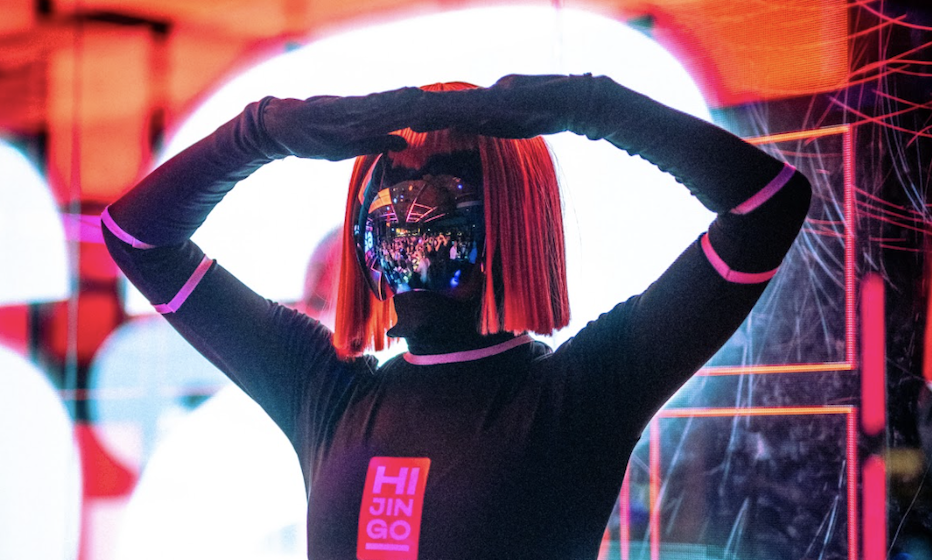 Person in futuristic attire with mirrored visor and orange wig in a travel-themed setting, with reflections of a crowd. Text on outfit reads &quot;HI JINGO.&quot;