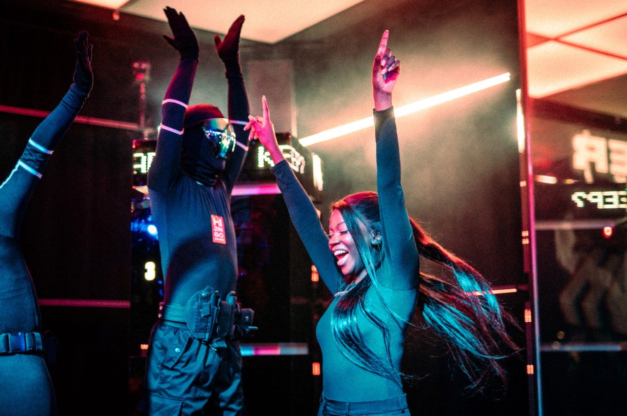 Three people excitedly raise their arms inside a high-tech travel-themed attraction