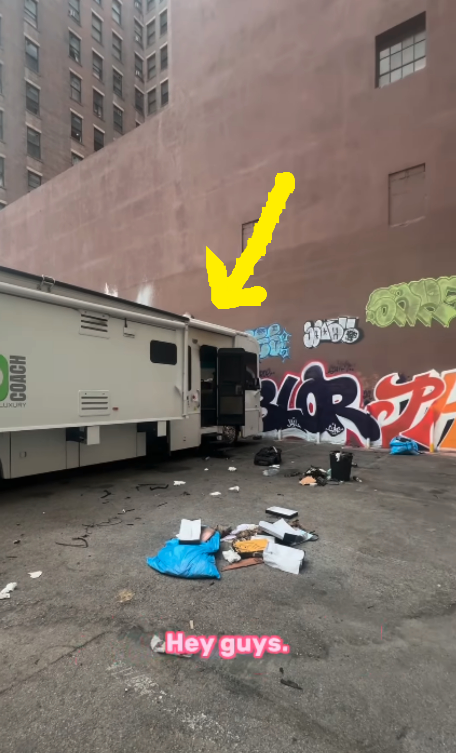 A parking lot with a camper van near a building with graffiti. Various items and trash are scattered on the ground. Text reads, "Hey guys."