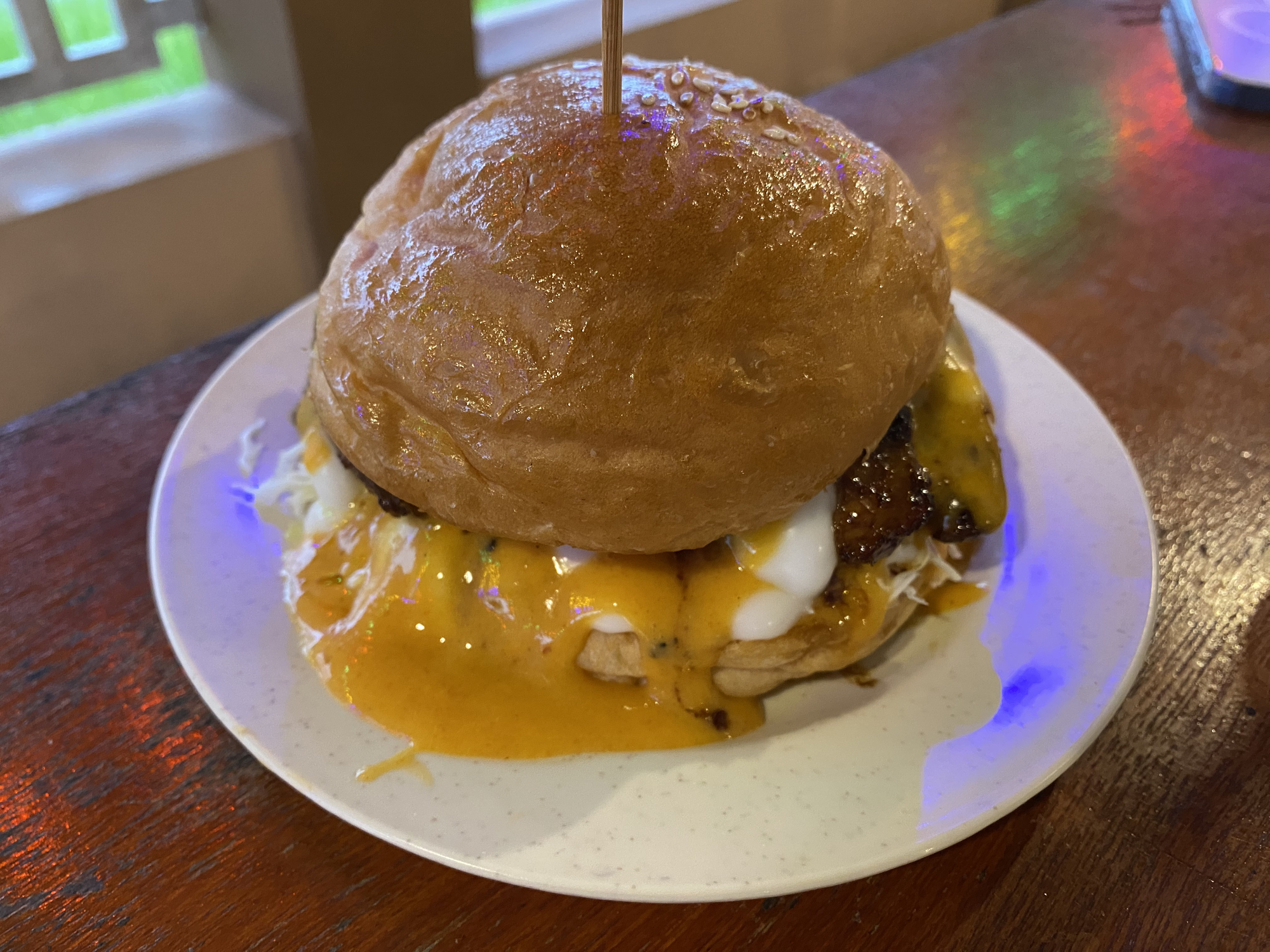 Close-up of a cheeseburger with melted cheese, lettuce, and sauce on a white plate. The background shows a window with a bit of greenery visible
