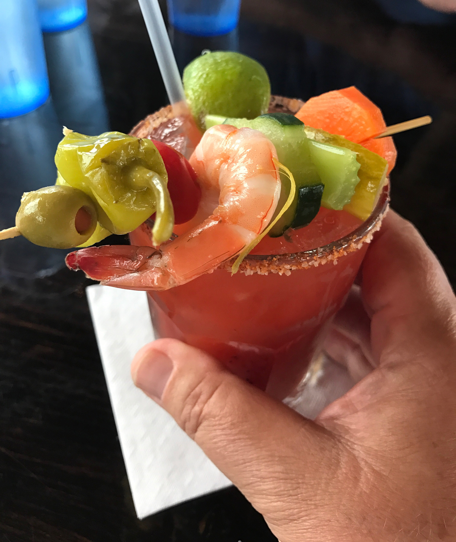 A hand holding a cocktail garnished with shrimp, olives, lime, cherry tomato, celery, and a pepper. The drink has a rim of salt or spices
