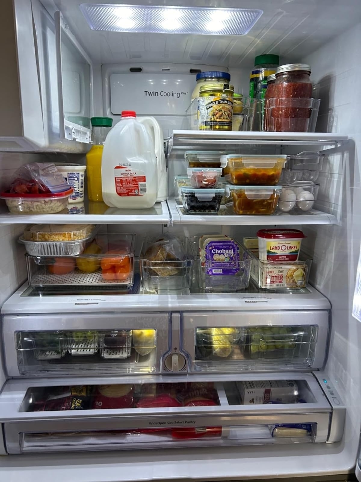 A well-organized refrigerator with shelves stocked with various groceries, including milk, juice, eggs, fruits, vegetables, and deli items
