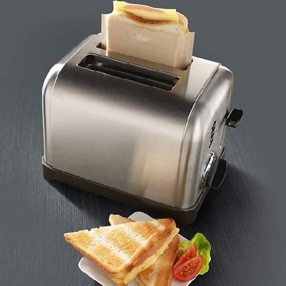 A stainless steel toaster with a sandwich in a heat-resistant bag being toasted. Below, a triangular toasted sandwich with cheese and tomato slices on a plate