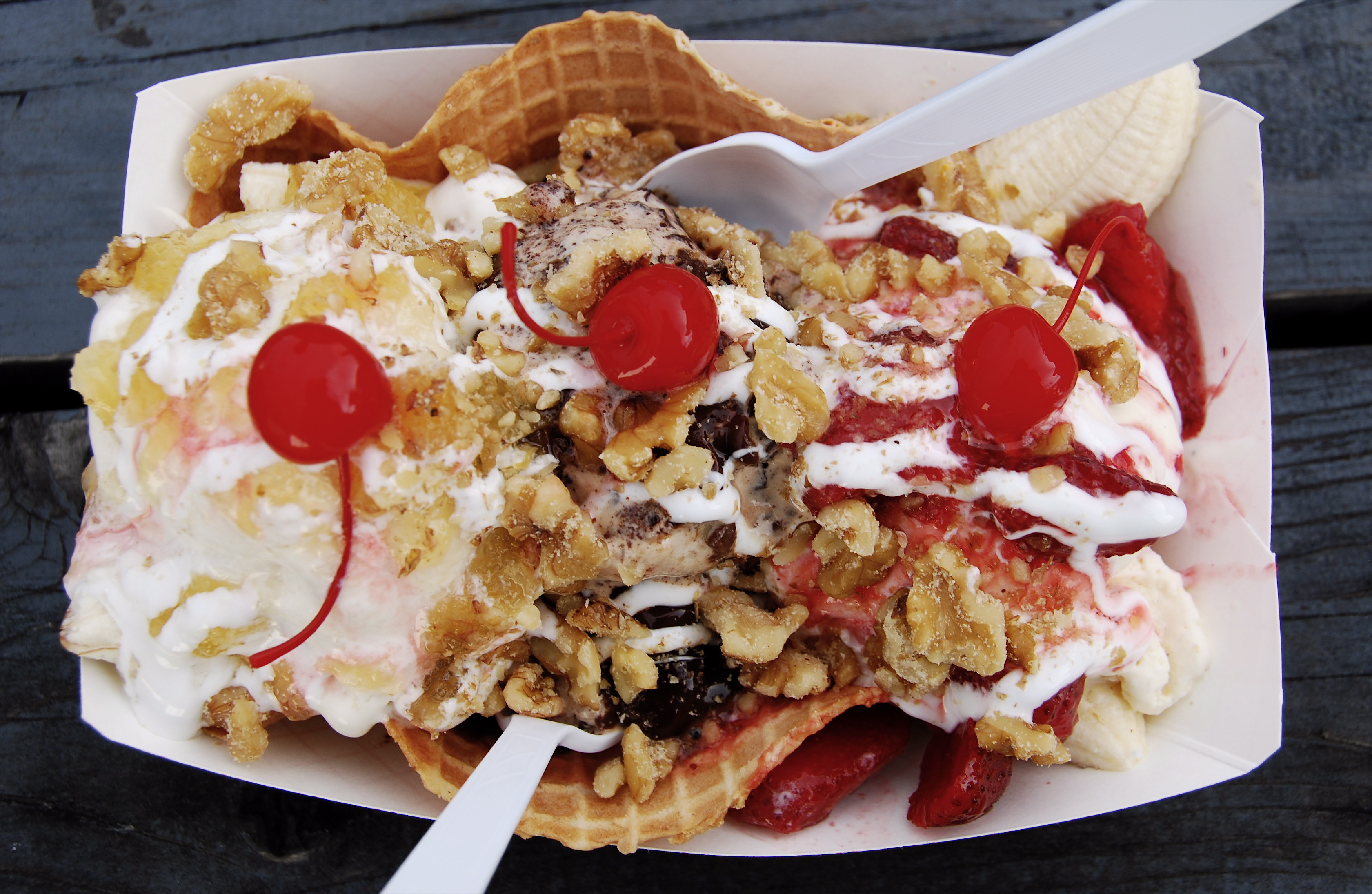 A close-up of a waffle bowl sundae topped with cherries, walnuts, ice cream, and whipped cream, served with two white spoons