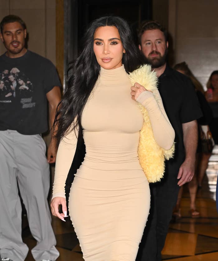 Kim Kardashian in a form-fitting dress, holding a fuzzy coat, walks with others in a public setting
