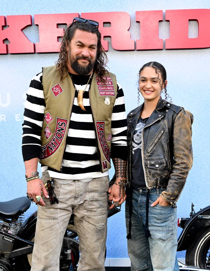 Jason Momoa and Lola Momoa on a red carpet, Jason wearing a striped shirt, vest, and cargo pants, and Lola in a leather jacket and jeans, holding hands
