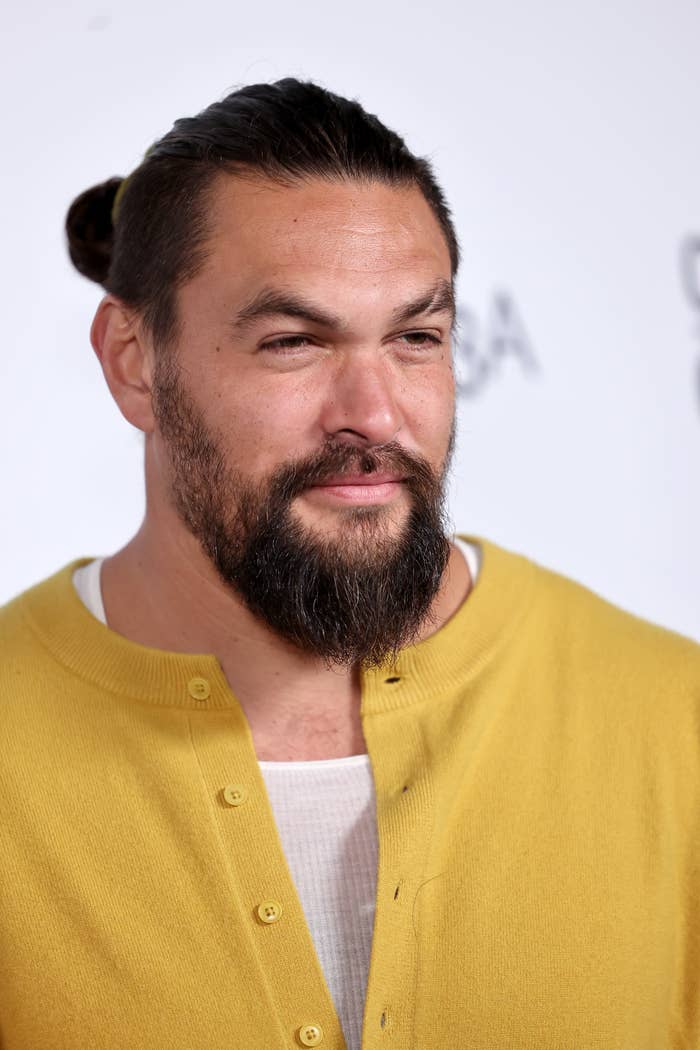 Jason Momoa on a red carpet, wearing a casual yellow cardigan over a white shirt