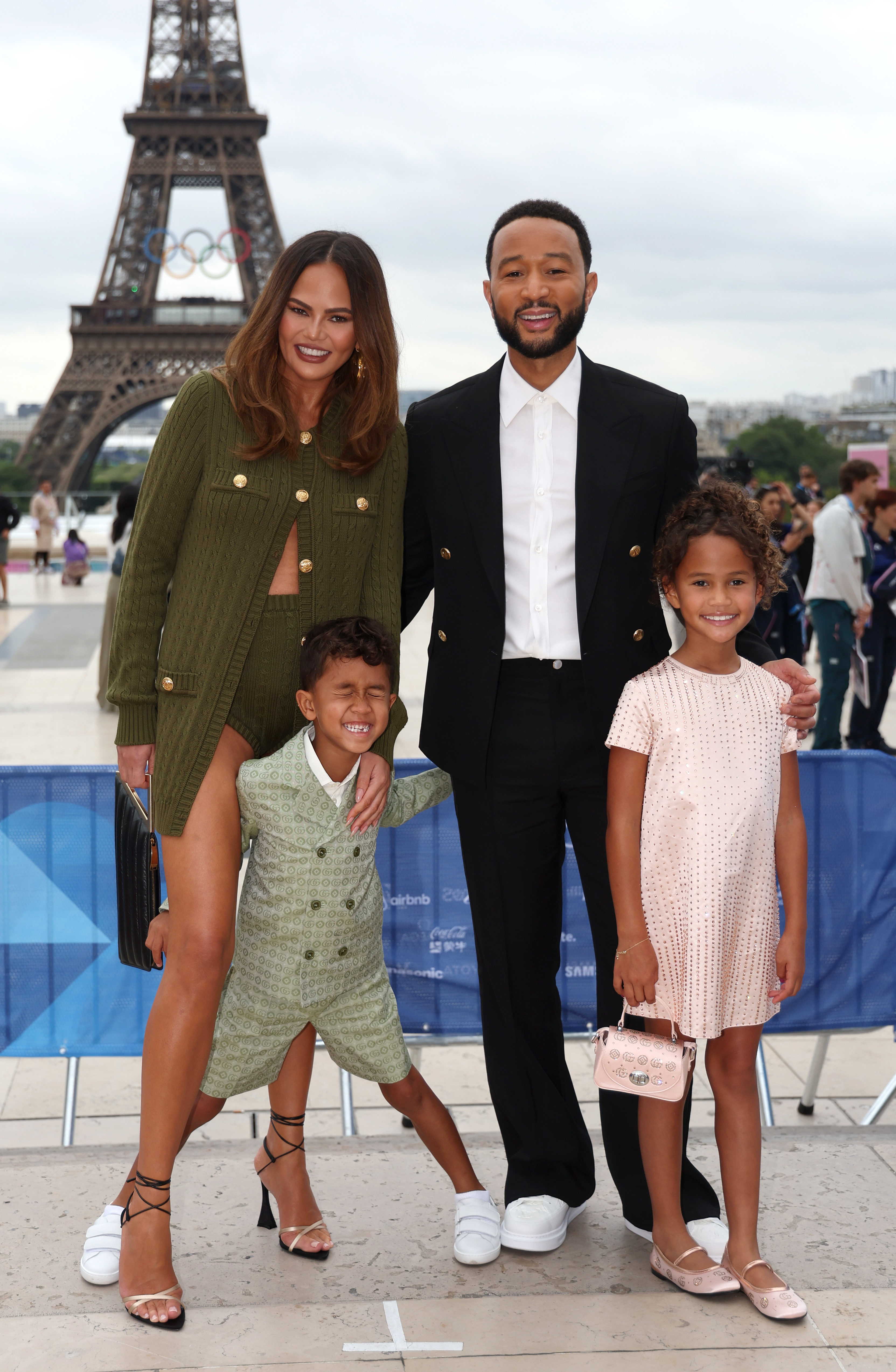 Chrissy Teigen and John Legend smile with their children, posing in stylish outfits in front of the Eiffel Tower