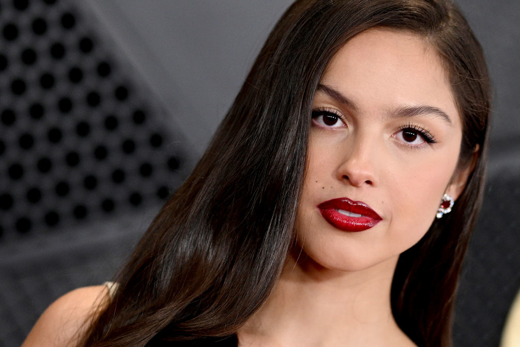 Olivia Rodrigo poses for a close-up photo with long hair styled to the side, wearing bold red lipstick and gemstone earrings