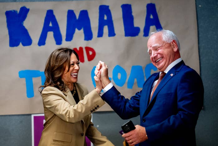 Kamala Harris and Tim Walz laughing and clasping hands in front of a sign that reads &quot;Kamala and Tim.&quot; Kamala wears a beige suit, and Tim wears a dark suit and tie