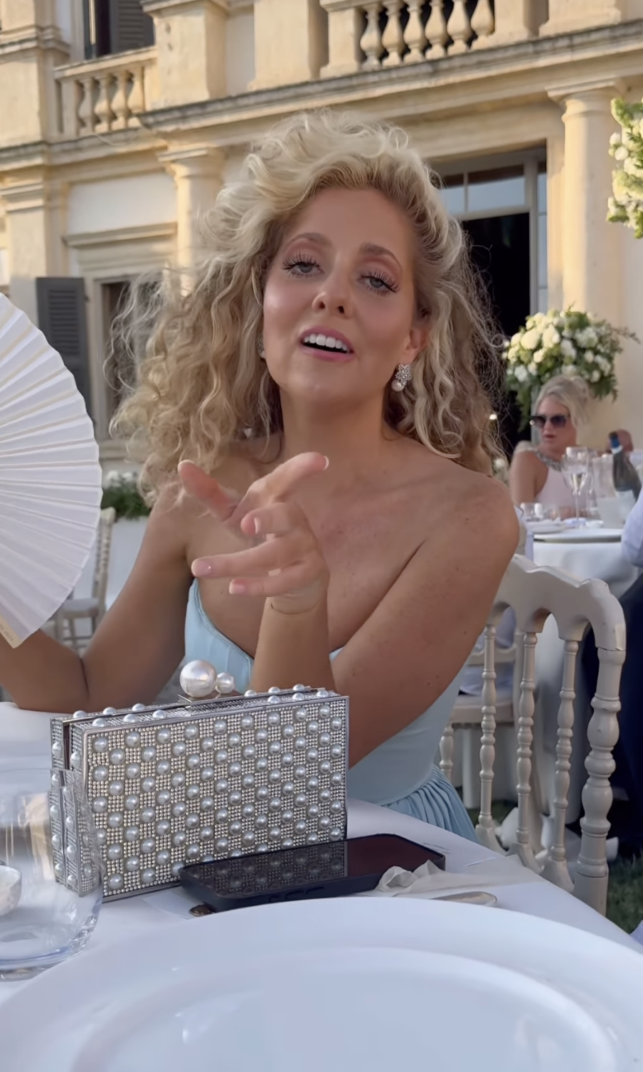 Roxy Horner sits at an outdoor event table, smiling with curly hair and holding a fan. She wears an elegant, strapless dress and is beside a sparkling clutch