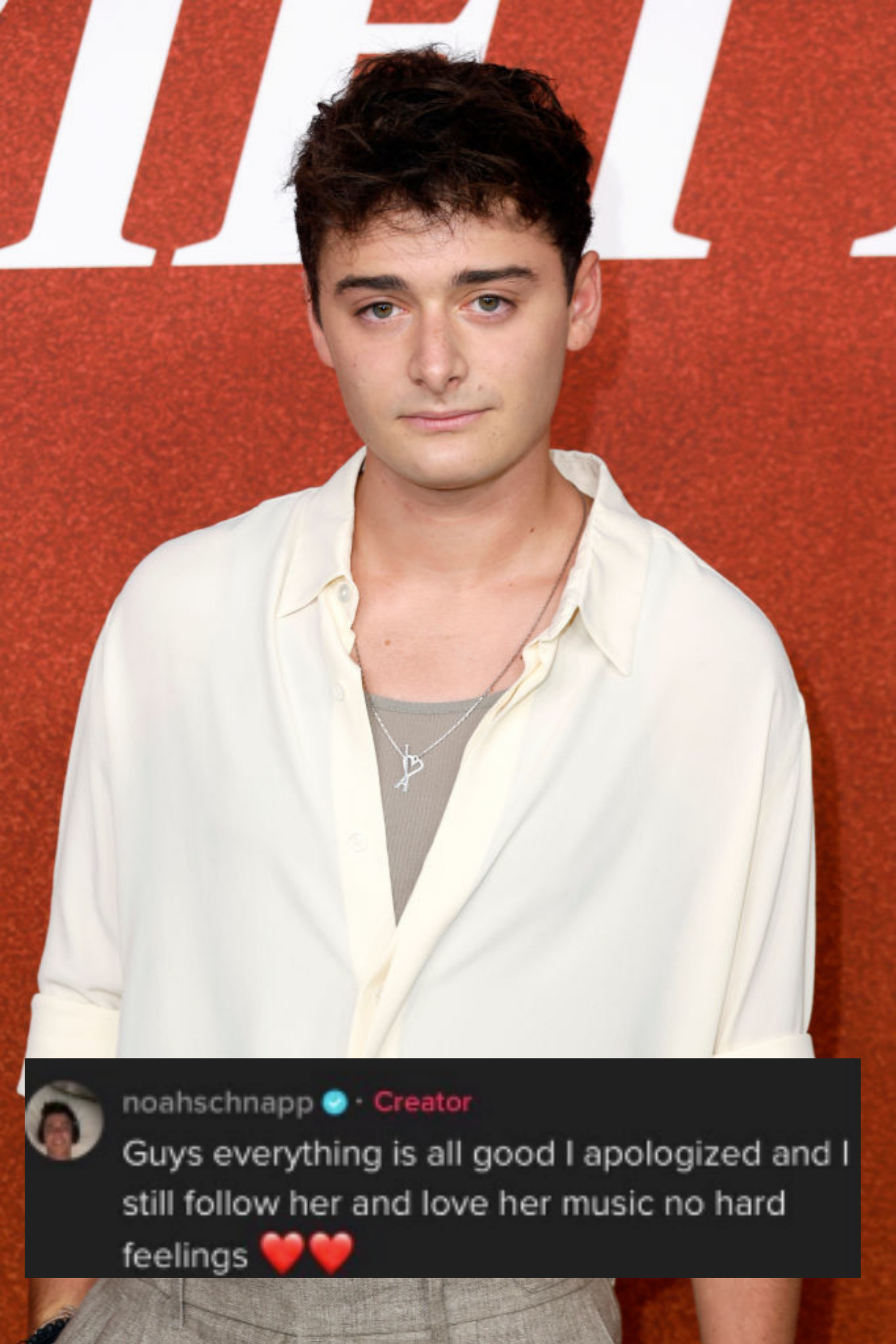 Noah Schnapp with short dark hair wears a loose white shirt over a light tank top and light grey pants, standing in front of a red background