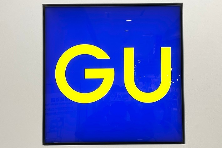 The image shows a square sign with blue background and large yellow letters &ldquo;GU&rdquo; in the center