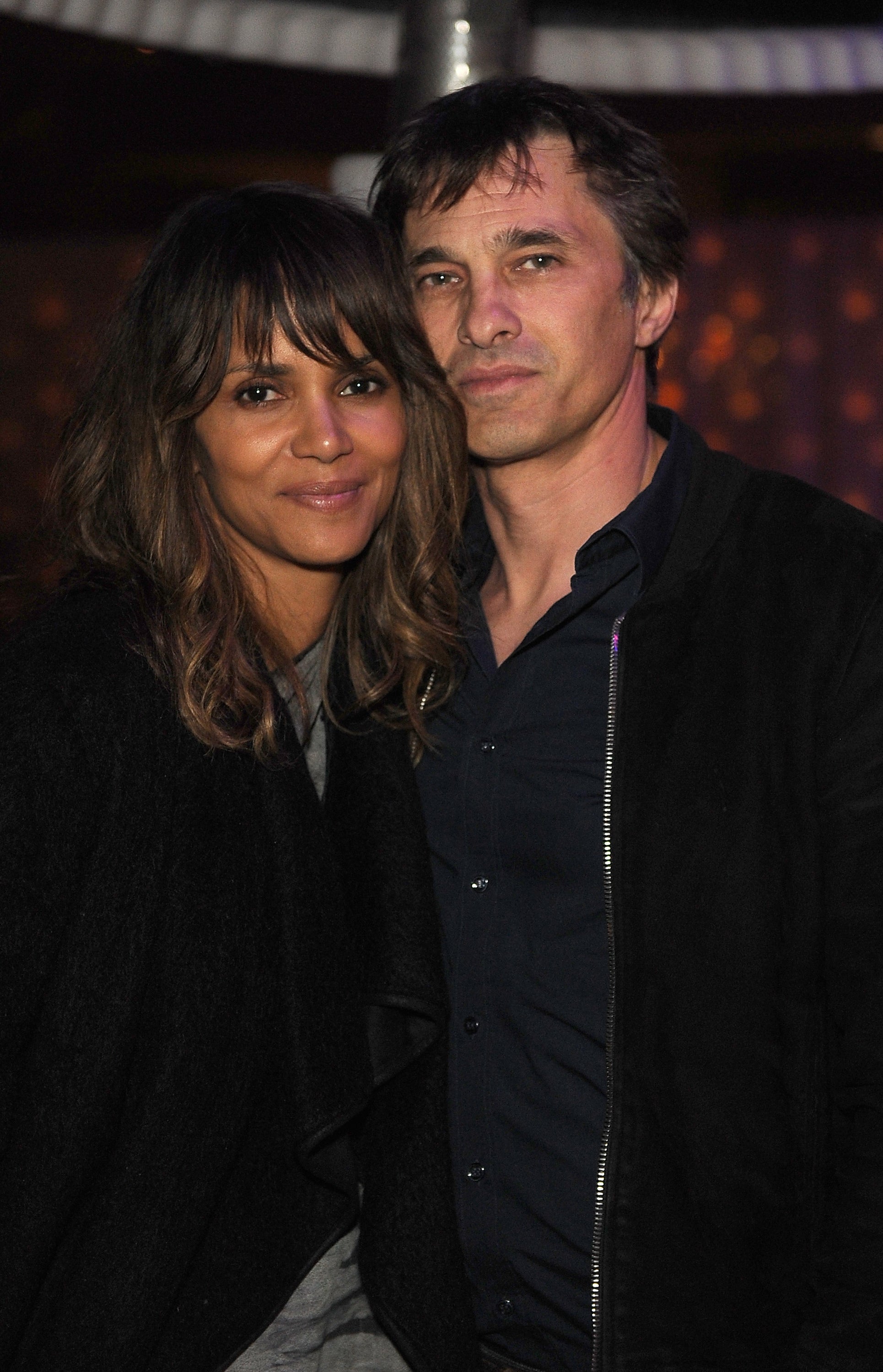 Halle Berry and Olivier Martinez stand closely together, smiling softly at the camera. Halle is wearing a casual outfit, and Olivier is in a dark jacket