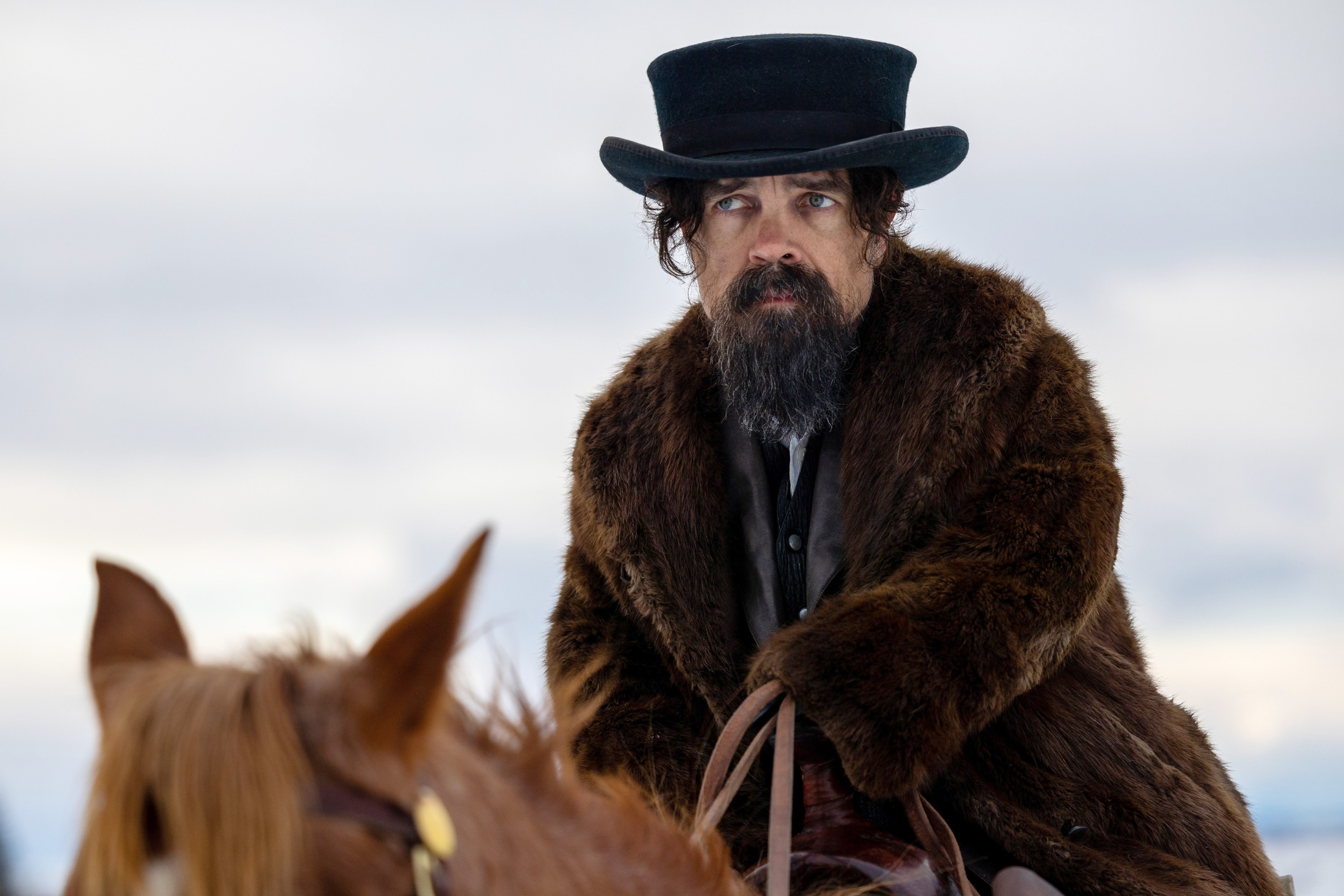 Peter Dinklage wearing a fur coat and a top hat, riding a horse in a winter landscape