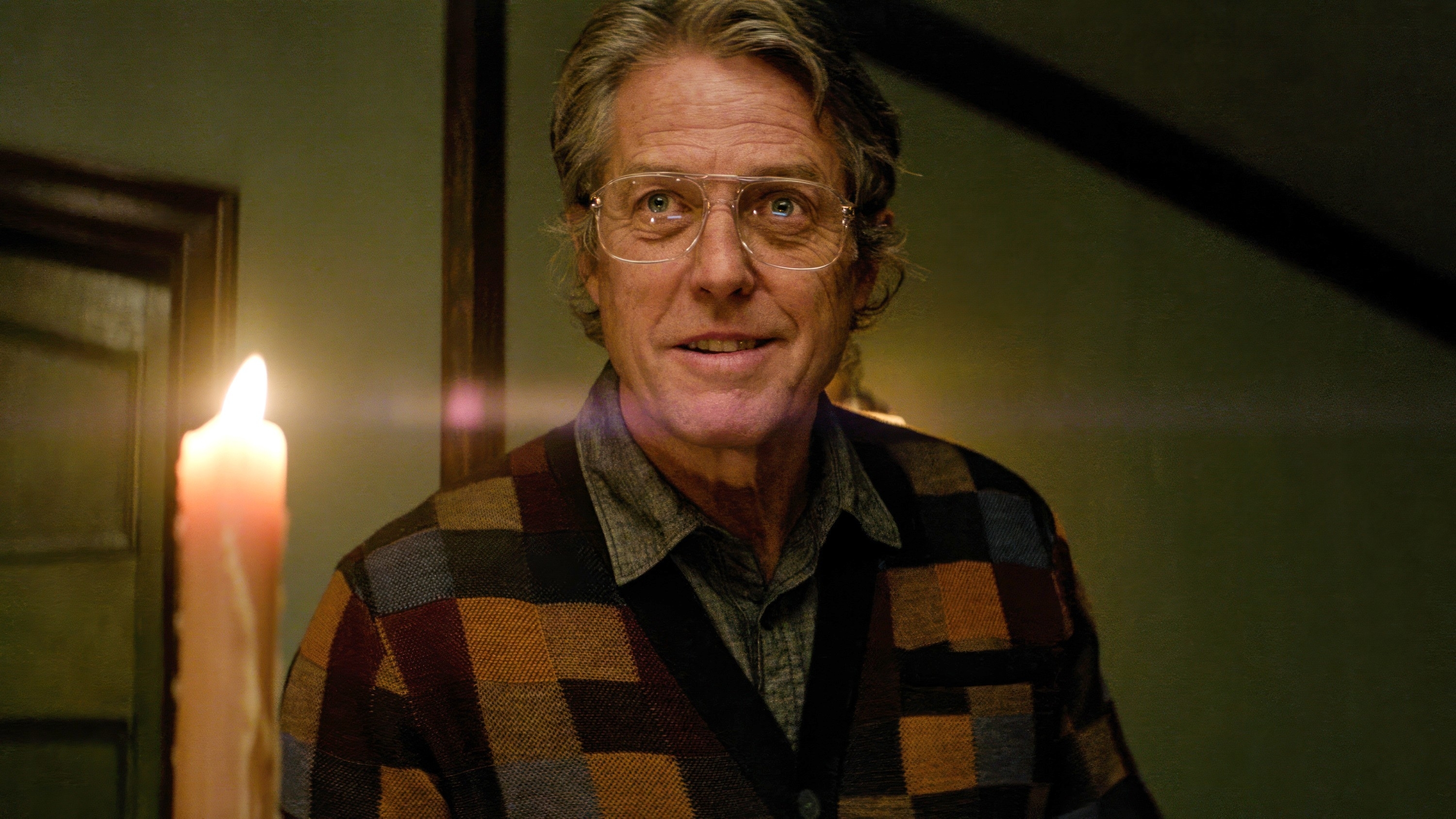 Hugh Grant smiling, wearing glasses and a multicolored checkered cardigan. There is a lit candle in the background