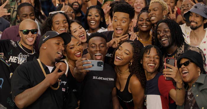 Large, diverse group of excited people taking a selfie, possibly cast members from TV or movie, with one person holding the phone. Others smile and pose