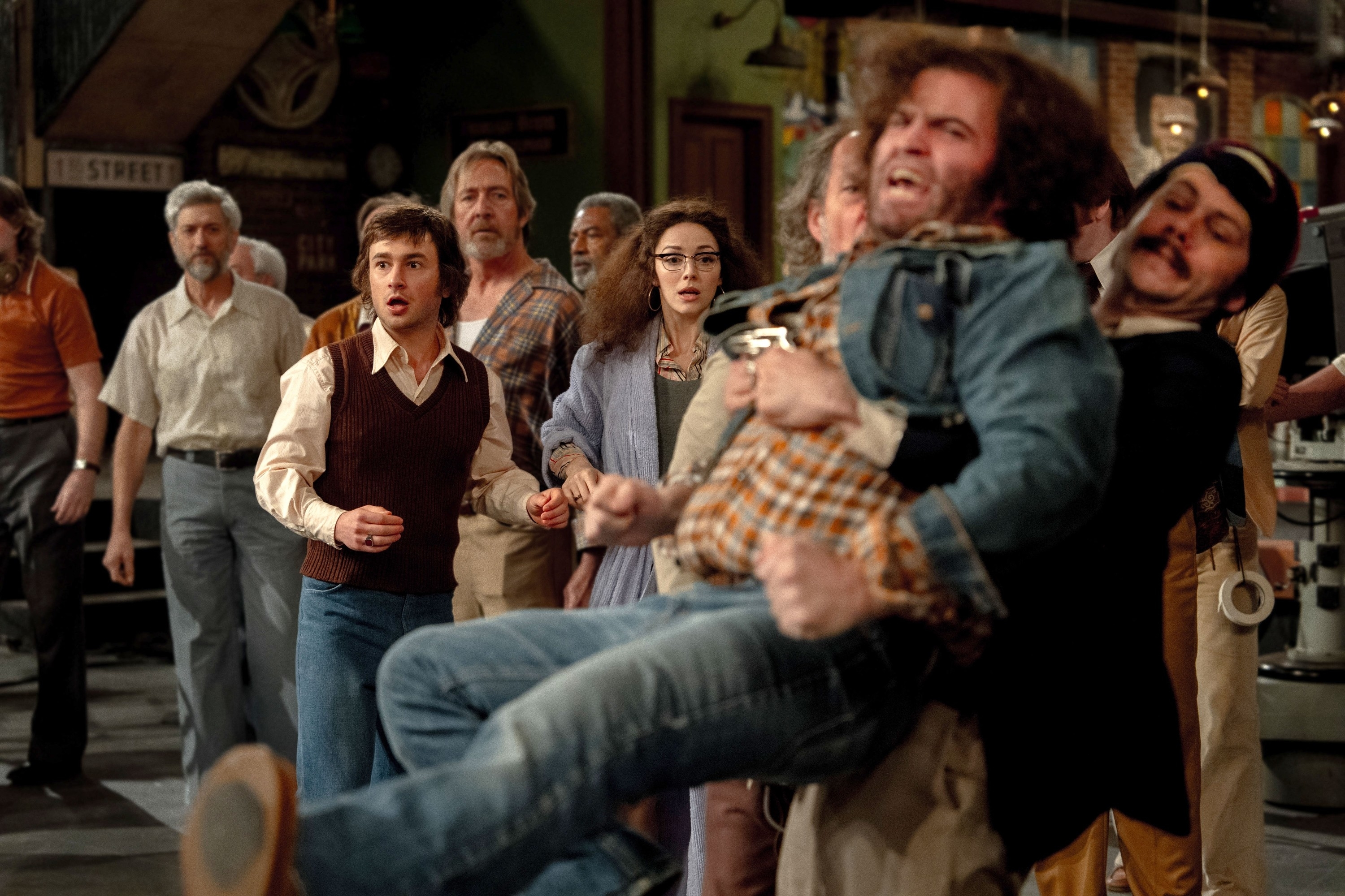 A chaotic scene with multiple people in 1970s attire. Central figures include Daniel Radcliffe, who appears shocked, and Paul Dano, who is frustrated
