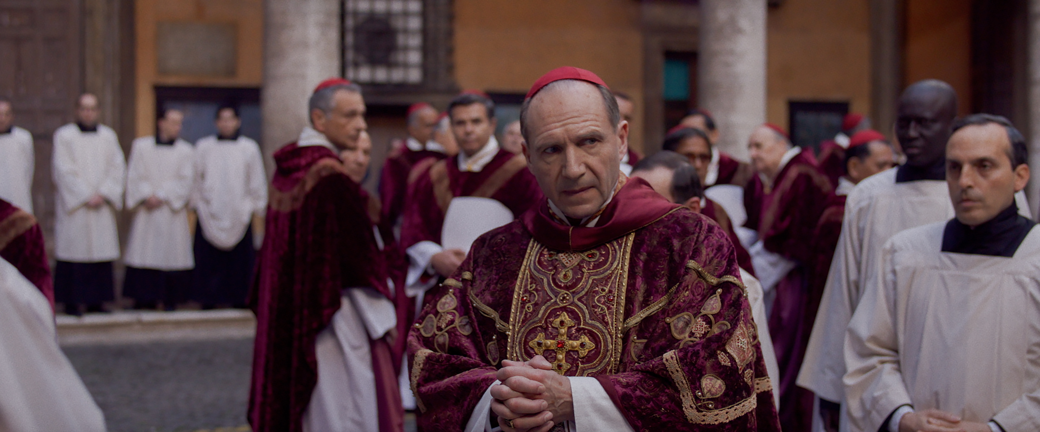 Group of clergy in ornate religious attire, including person in focus wearing a gold and red embroidered robe and a red zucchetto, surrounded by others in similar robes