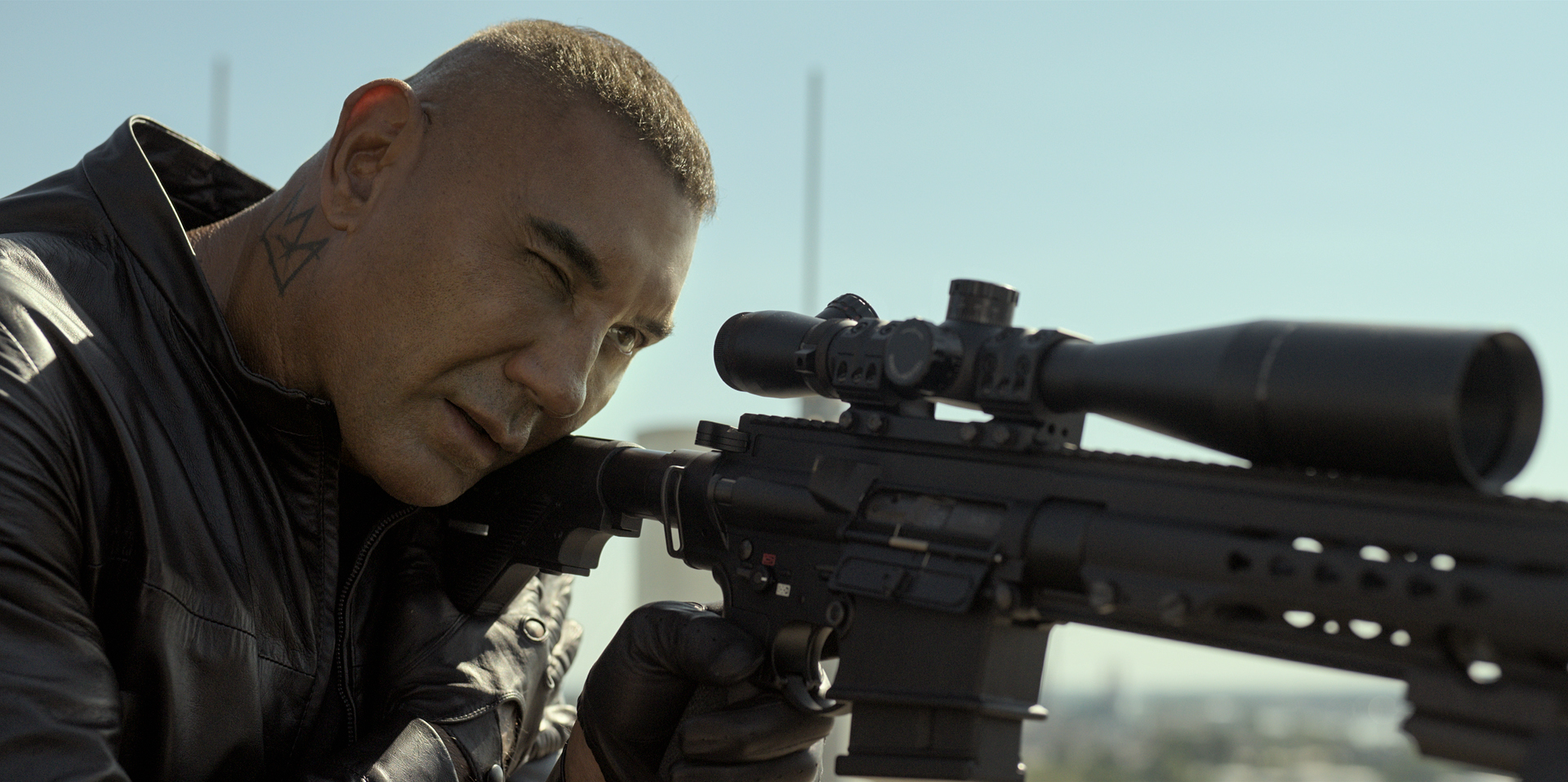 Temuera Morrison aiming through the scope of a sniper rifle, wearing a black jacket and gloves