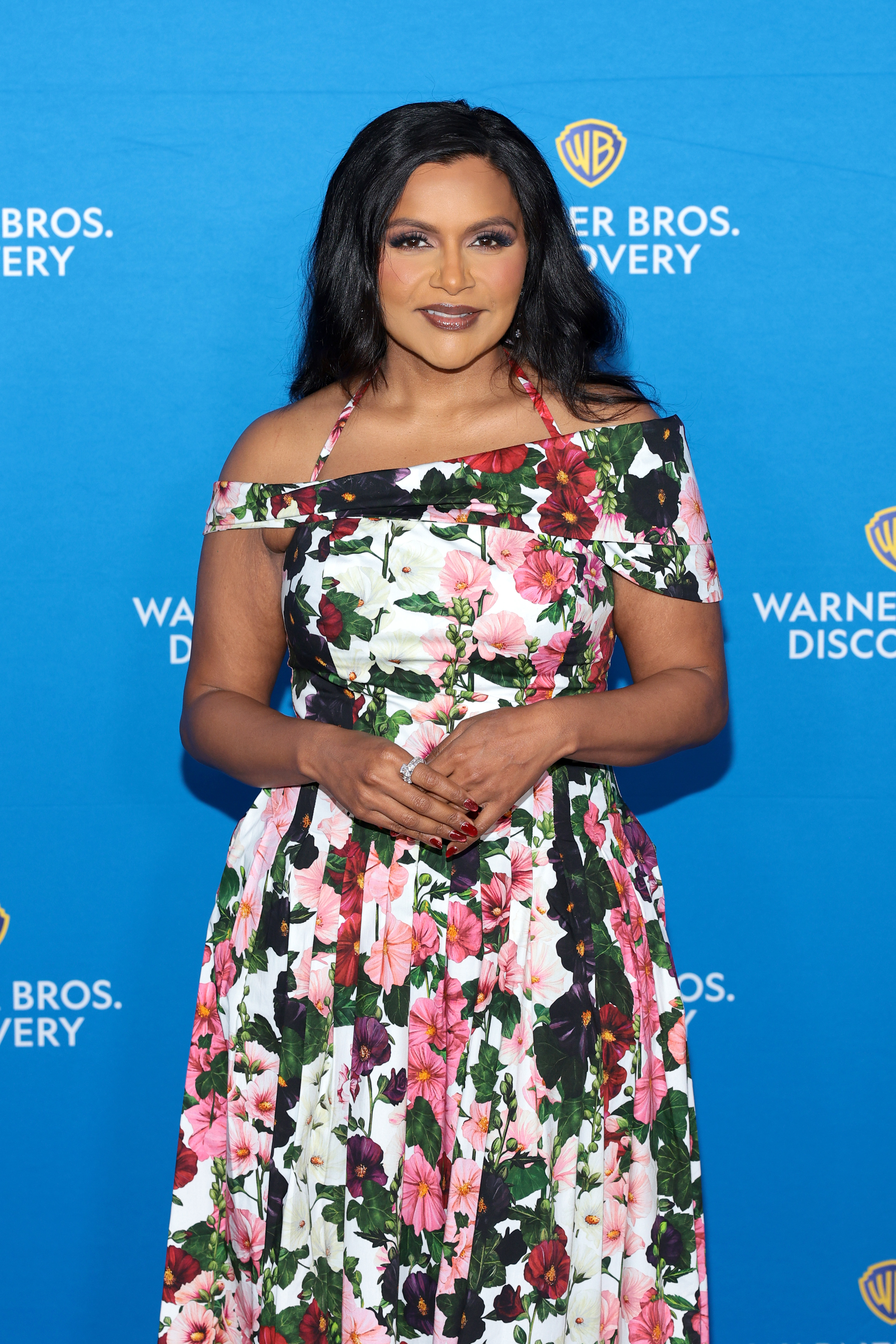 Mindy Kaling at a Warner Bros. Discovery event, wearing a floral dress with an off-shoulder design, standing in front of a blue background