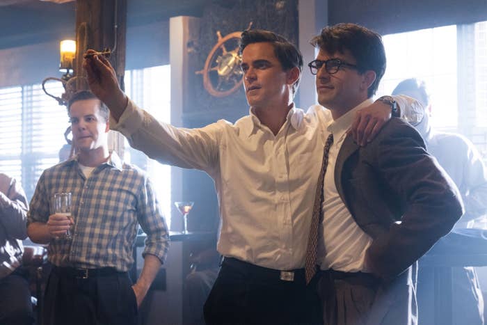 Men dressed in mid-20th-century attire at a bar; one man has his arm around another. Scene from a period film or TV show featuring actors