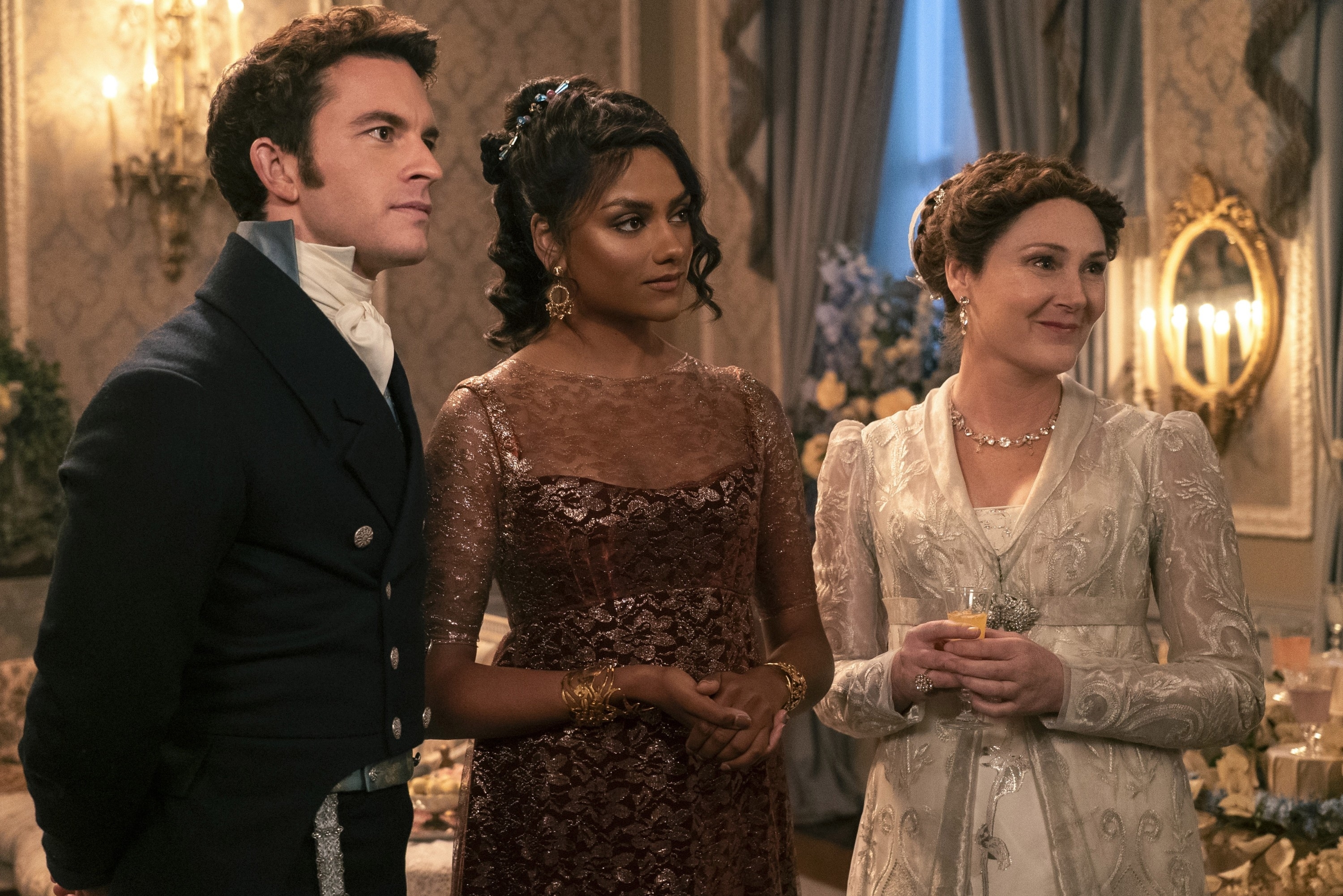 Jonathan Bailey, Simone Ashley, and Polly Walker standing together at a formal event, dressed in period costume attire