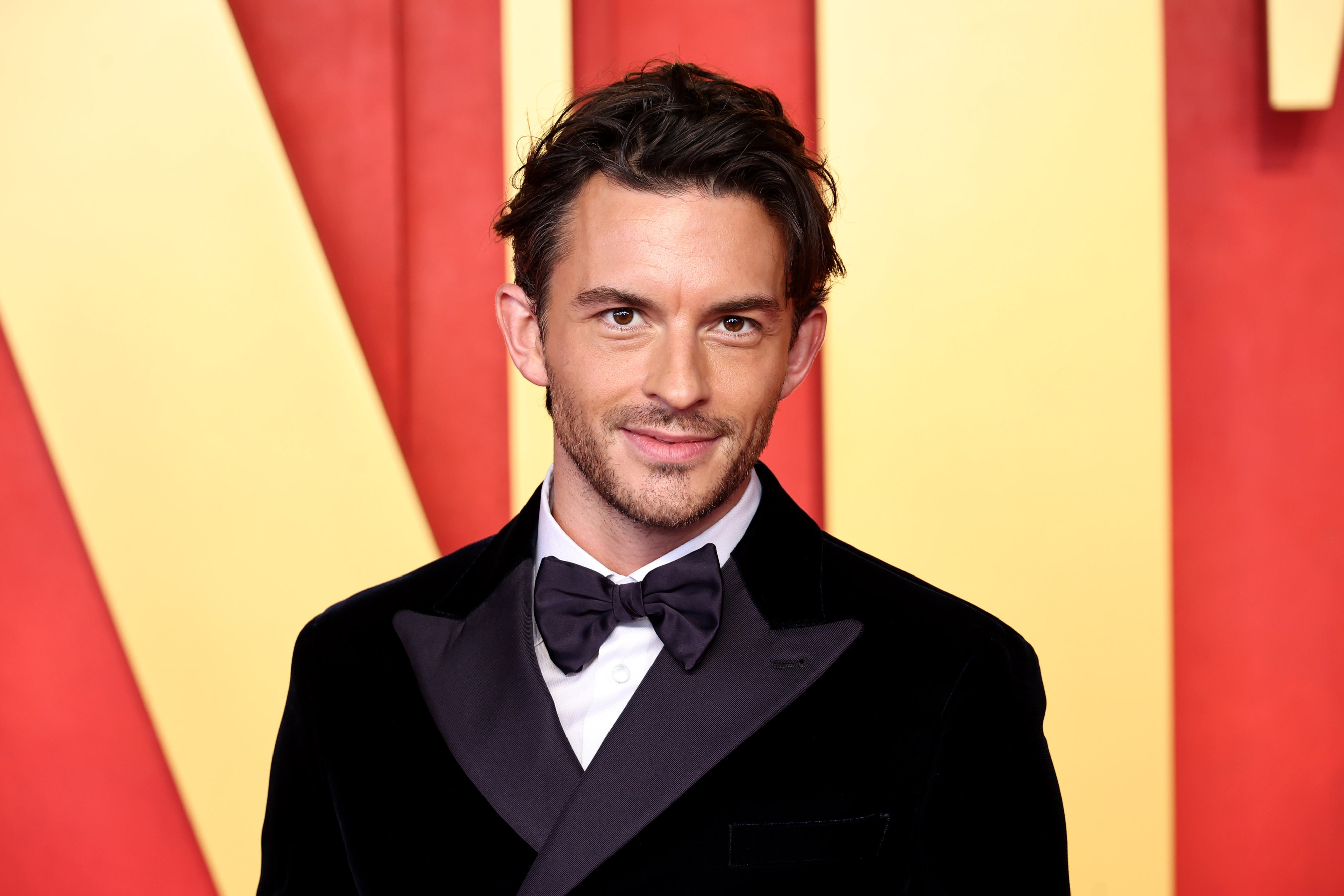 Jonathan Bailey poses in a black tuxedo with a bow tie on a red carpet