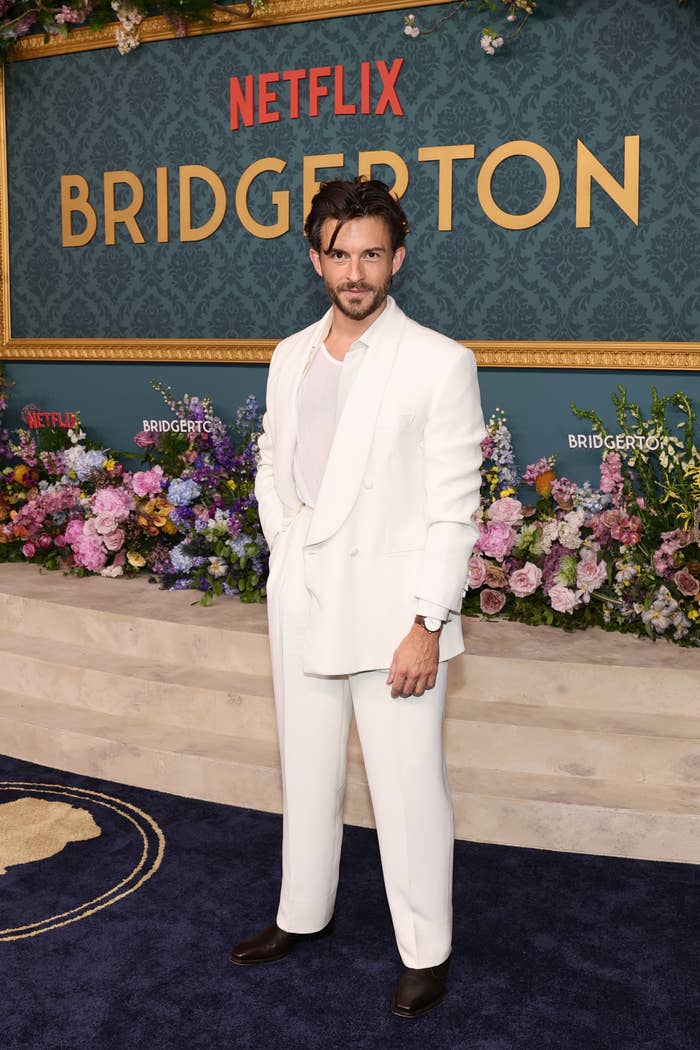 Jonathan Bailey wearing a white suit stands on the Netflix "Bridgerton" event's red carpet backdrop, which is adorned with floral arrangements