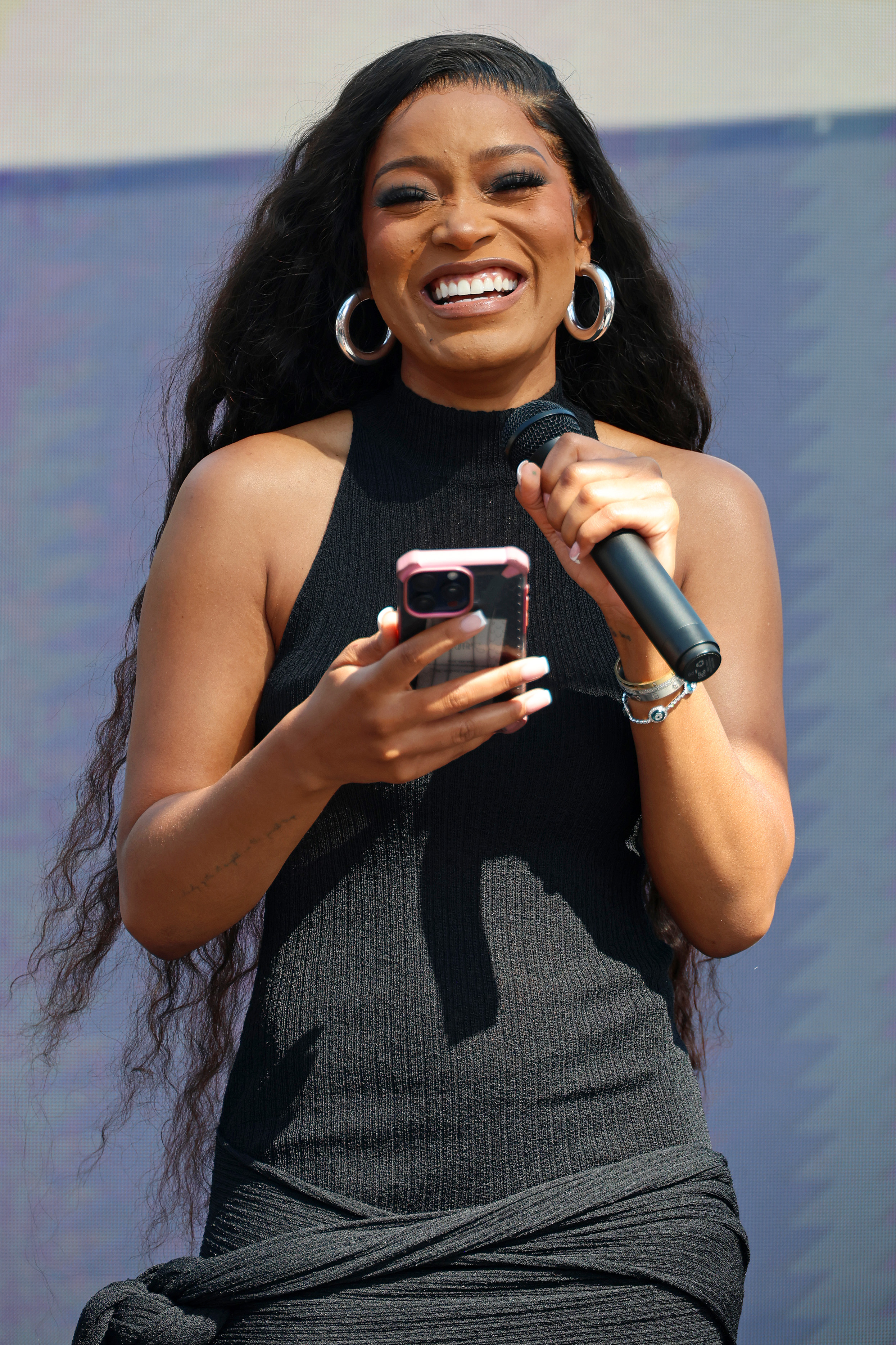 Keke Palmer smiles and holds a phone and a microphone while wearing a black halter dress