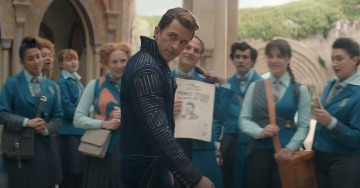 Jonathan Bailey, in a dark suit with intricate designs, holds up a newspaper surrounded by eight students in blue uniforms near a stone building