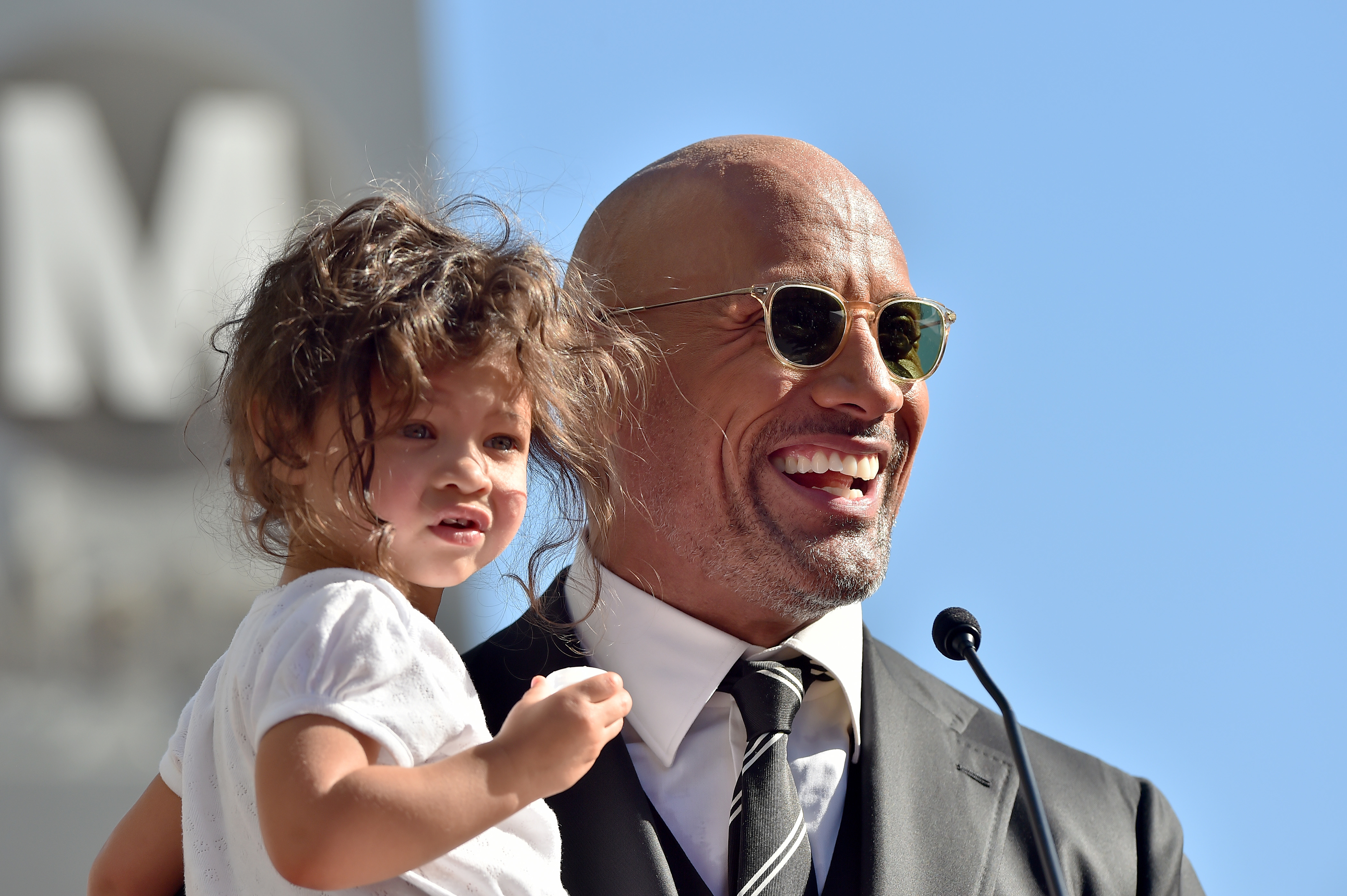 Dwayne Johnson holding his daughter at an event, smiling and wearing a suit with sunglasses. His daughter is wearing a casual white outfit