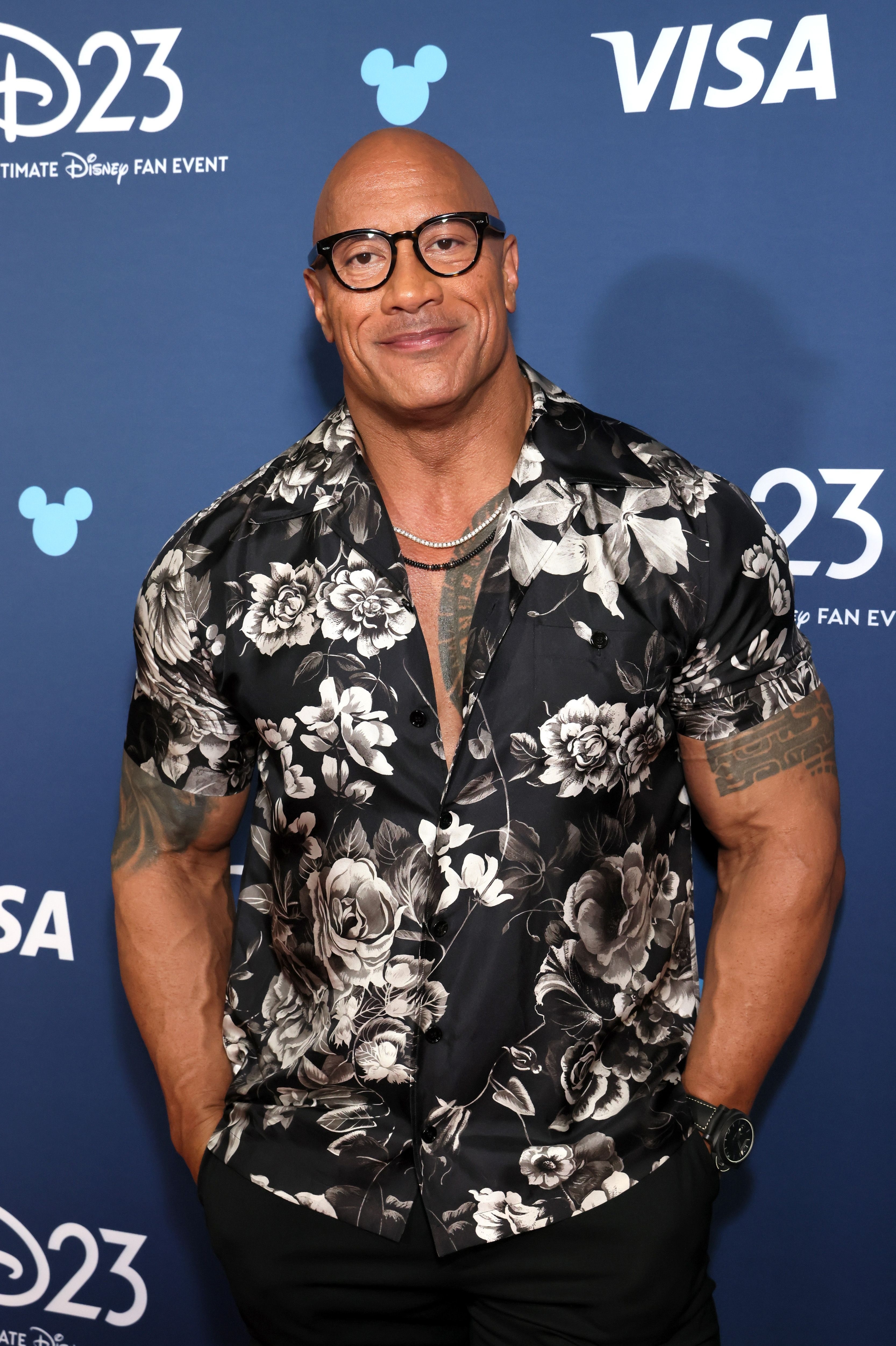Dwayne Johnson at the D23 Disney event, wearing a floral patterned short sleeve shirt, glasses, and a necklace
