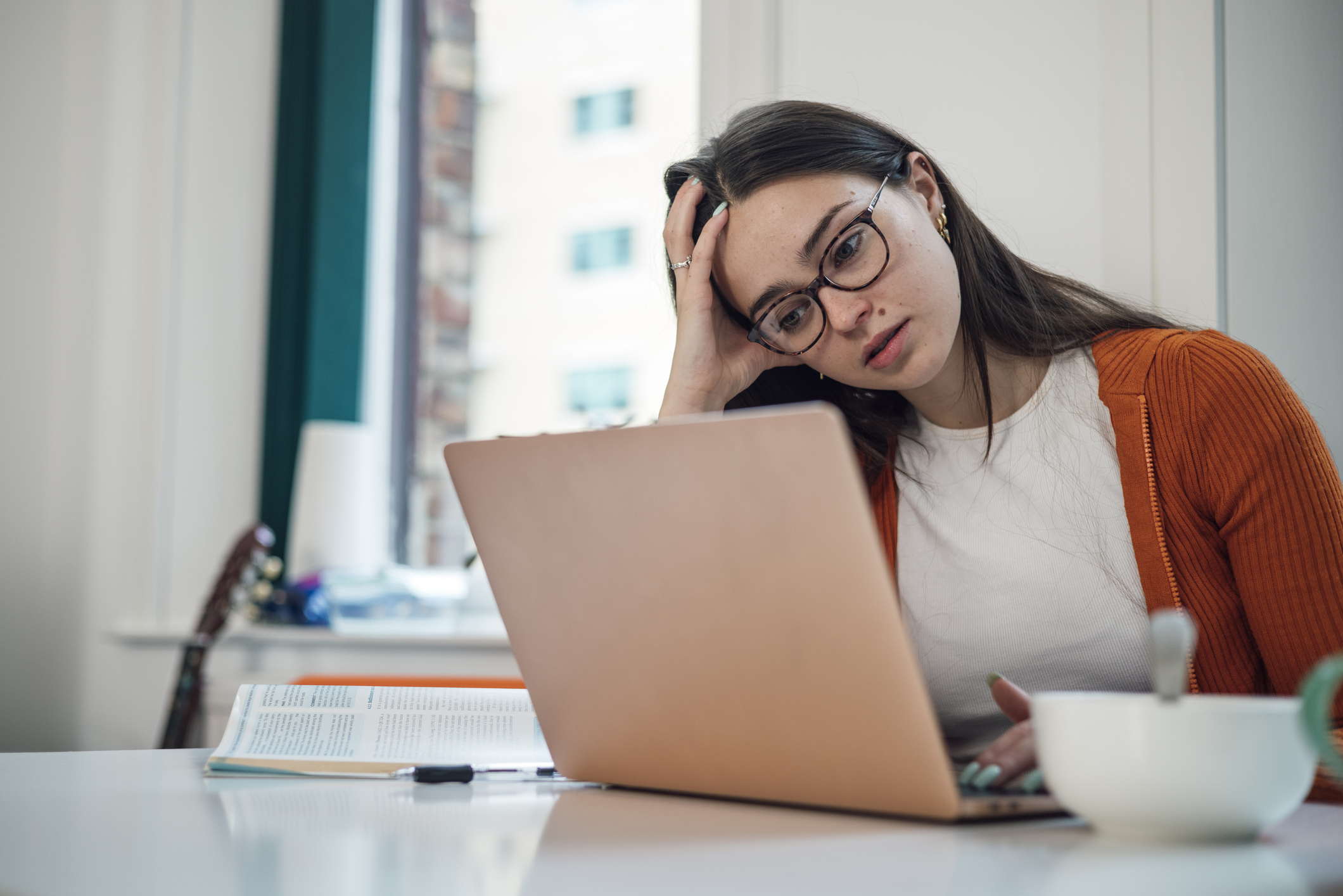 Woman sitting at a desk, holding her head and looking stressed while working on a laptop. There's an open book and cup next to her